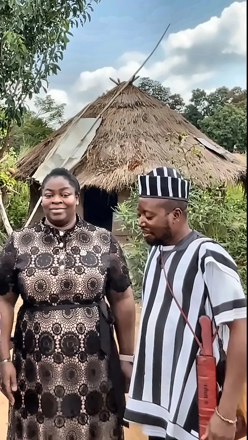 A woman and a man in A’nger cloth in front of a thatched hut