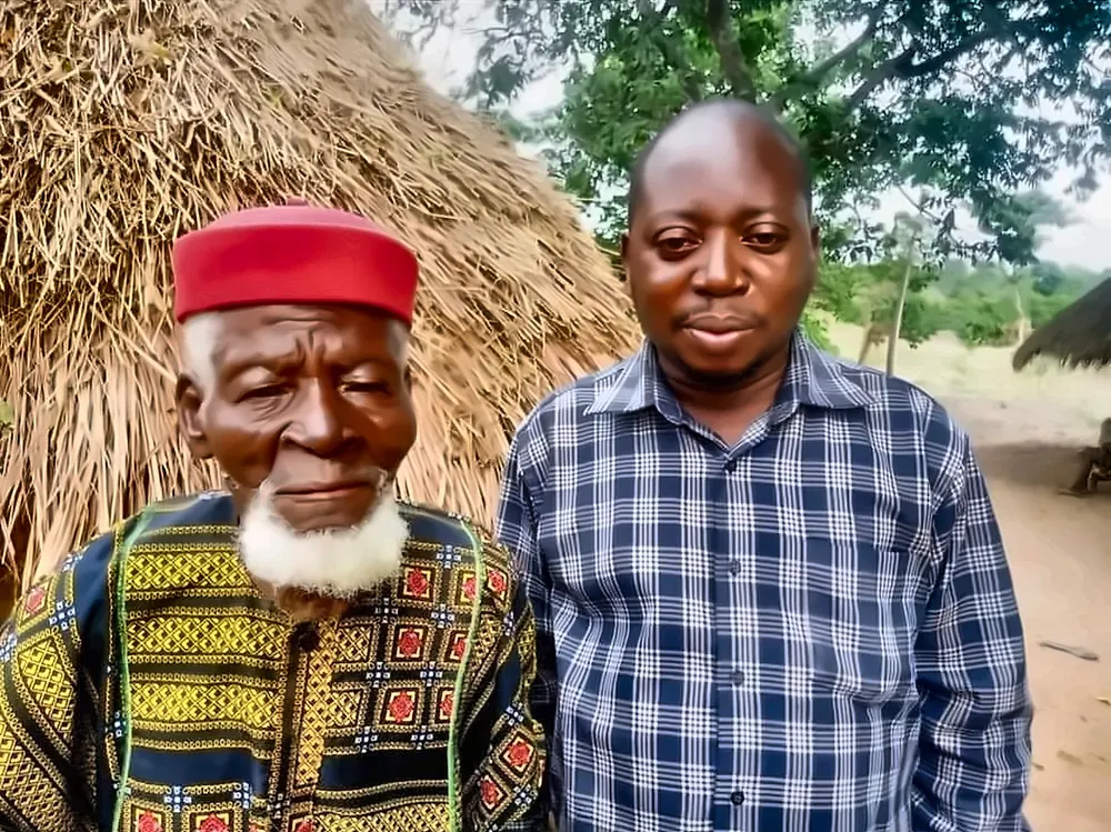 An elder in a red cap standing with a younger man in front of a thatched hut