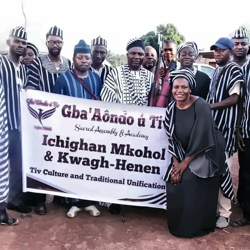 A woman kneels before the Ichighan Mkohol & Kwagh-Henen banner, surrounded by members in A’nger cloth